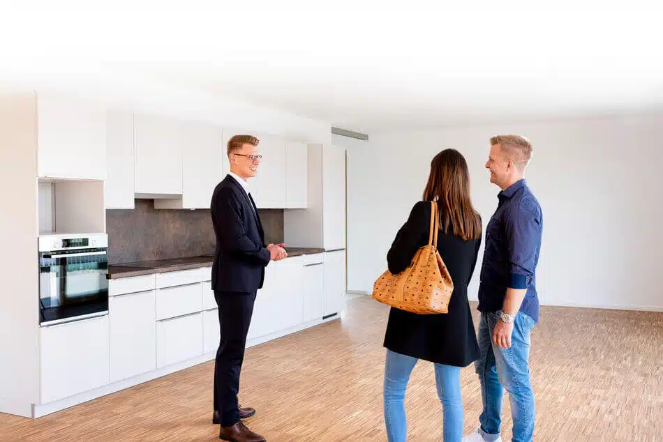 Entwicklung der Hauspreise in Deutschland in den Jahren von 2000 bis 2022 Ein Immobilienmakler in einem Anzug zeigt einem lächelnden Paar eine moderne, leere Küche. Das Paar steht zusammen, dem Makler zugewandt, in einem geräumigen, gut beleuchteten Raum mit Holzfußboden.