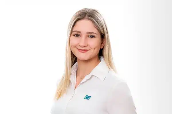 Eine junge Frau mit glattem, blondem Haar, die eine weiße Bluse mit einem kleinen blaugrünen Logo trägt, lächelt und steht vor einem weißen Hintergrund.
