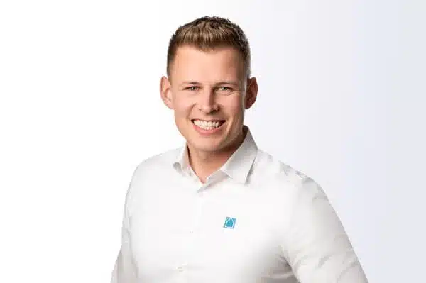Ein junger Mann mit kurzem blondem Haar lächelt in die Kamera. Er trägt ein weißes Button-up-Hemd mit einem kleinen blau-grünen Logo auf der Brust und steht vor einem Hintergrund mit weißem bis hellgrauem Farbverlauf.