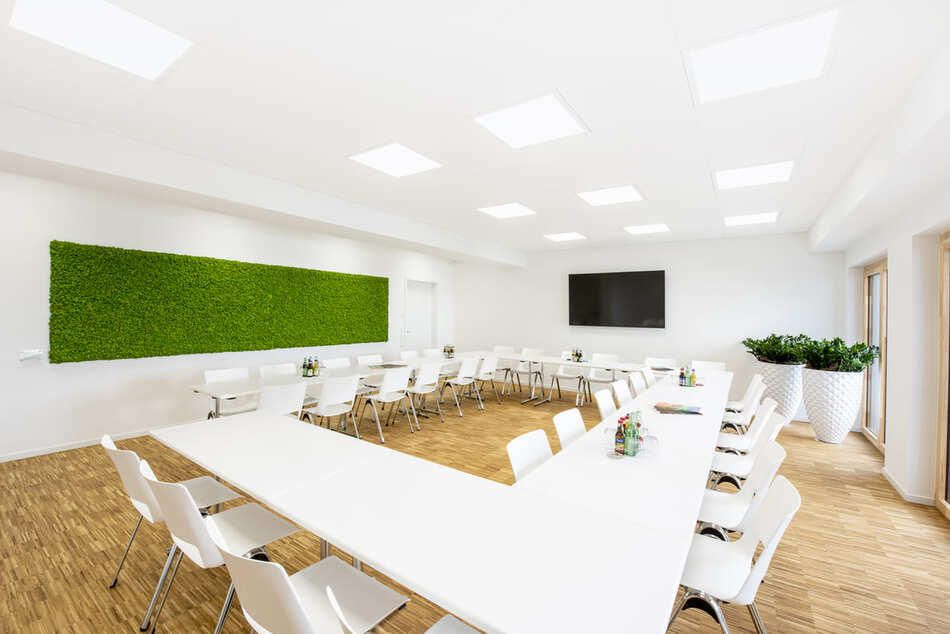 Ein heller, moderner Konferenzraum mit weißen Stühlen und Tischen, die in U-Form angeordnet sind. Der Raum verfügt über eine große grüne Pflanzenwand, einen Holzboden, Topfpflanzen und einen an der Wand befestigten Flachbildfernseher.