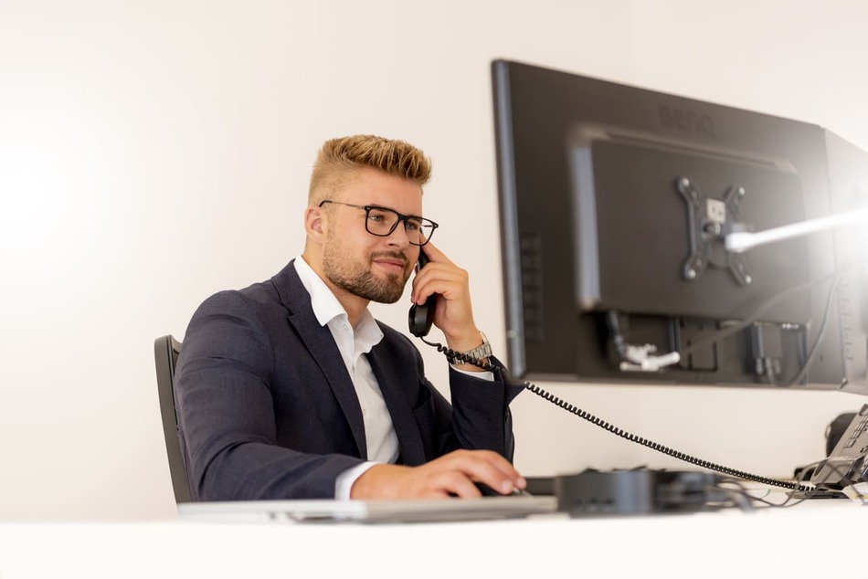 Köhl-Jetter Impressionen Ein Mann mit blondem Haar und Brille, im Anzug und am Schreibtisch sitzend, telefoniert, während er auf einen Computerbildschirm schaut.