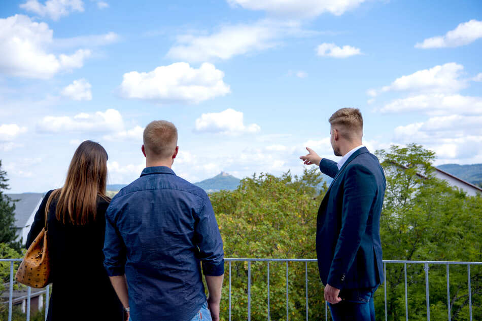Köhl-Jetter Impressionen Drei Personen stehen im Freien vor einer malerischen Landschaft; eine Person im Anzug zeigt auf ferne Hügel und einen blauen Himmel mit Wolken. Im Hintergrund sind Bäume und Dächer zu sehen.