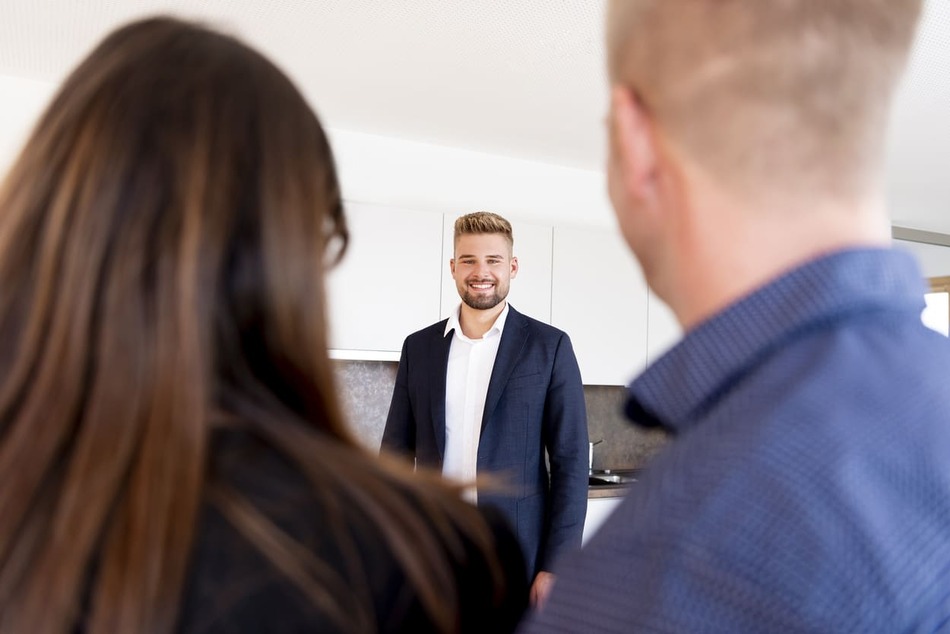 Köhl-Jetter Impressionen Ein lächelnder Mann im Anzug steht in einer hellen, modernen Küche und blickt auf ein Paar, das von hinten gesehen wird, was auf ein Gespräch oder eine Immobilienbesichtigung hindeutet.