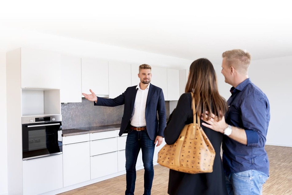Ein Immobilienmakler in einem Anzug zeigt einem Paar eine moderne Küche. Das Paar steht zusammen, wobei die Frau eine braune Handtasche trägt, während der Makler in Richtung der Küchenschränke und -geräte gestikuliert.