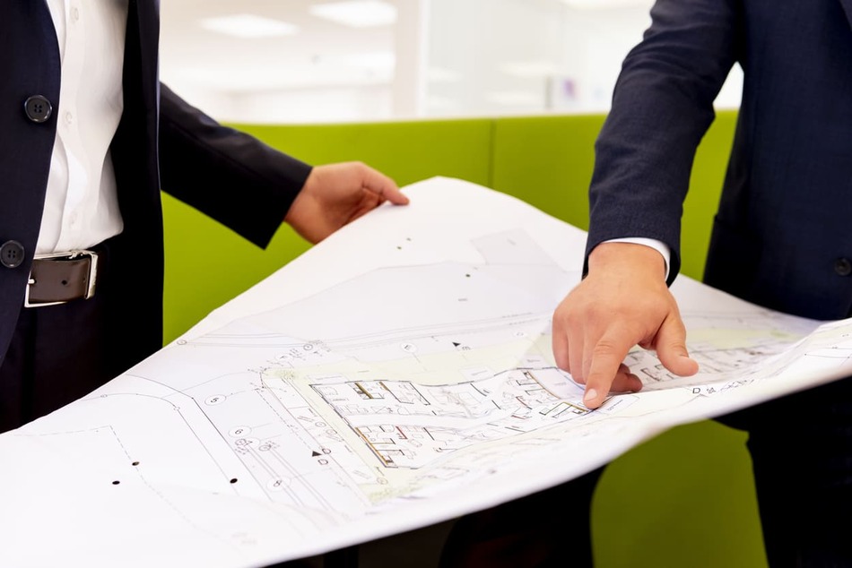 Zwei Männer in Geschäftskleidung stehen in einem hellen Büro und betrachten und besprechen einen großen Bauplan, der zwischen ihnen ausgebreitet ist. Ein Mann zeigt auf einen Ausschnitt der Blaupause, während der andere aufmerksam zuhört.