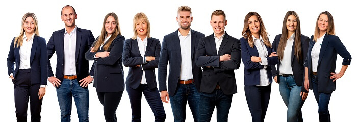 Köhl-Jetter Immobilien GmbH & Köhl-Jetter Hausverwaltungen GmbH & Co. KG Eine Gruppe von zehn jungen Erwachsenen, Männern und Frauen, in legerer Geschäftskleidung mit Blazern und Jeans, die in einer Reihe stehen und selbstbewusst vor einem weißen Hintergrund lächeln.