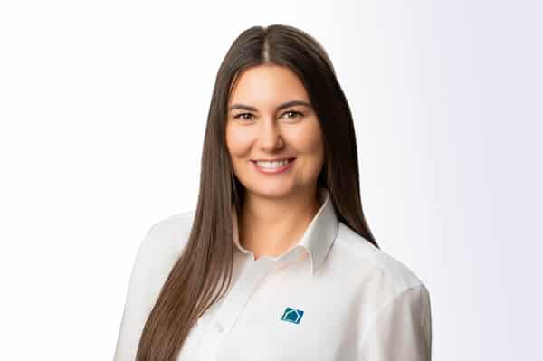 Eine Frau mit dunklem Haar, die ein weißes Button-up-Shirt mit einem blauen quadratischen Logo trägt, steht und lächelt in die Kamera vor einem weißen Hintergrund.