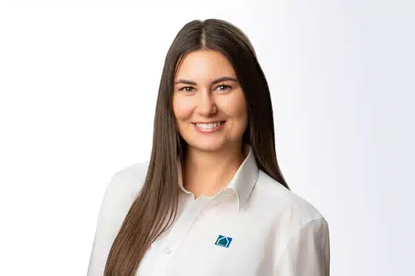 Eine Frau mit dunklem Haar, die ein weißes Button-up-Shirt mit einem blauen quadratischen Logo trägt, steht und lächelt in die Kamera vor einem weißen Hintergrund.