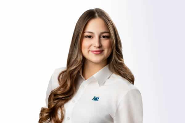 Eine Frau mit braunem Haar, die ein weißes Button-up-Shirt mit einem blauen quadratischen Logo trägt, steht und lächelt in die Kamera vor einem weißen Hintergrund.