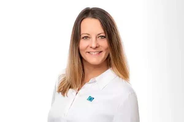 Eine Frau mit glattem, schulterlangem braunem Haar lächelt, während sie ein weißes Hemd mit einem blau-grünen Logo trägt und vor einem schlichten weißen Hintergrund posiert.