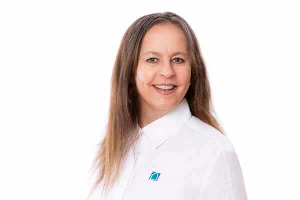 Eine Frau mit langen braunen Haaren lächelt in die Kamera. Sie trägt ein weißes Kragenhemd mit einem blau-grünen Logo und steht vor einem schlichten weißen Hintergrund.