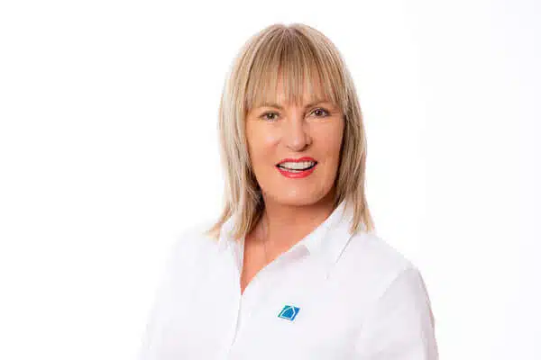 Eine Frau mit glattem blondem Haar, die ein weißes Button-up-Shirt mit einem blauen quadratischen Logo trägt, steht und lächelt in die Kamera vor einem weißen Hintergrund.