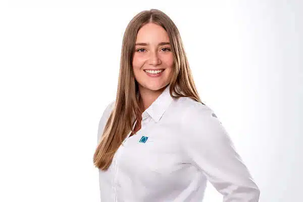 Eine Frau mit langem glattem Haar, die ein weißes Button-up-Hemd mit einem kleinen blauen Logo trägt, lächelt in die Kamera vor einem schlichten weißen Hintergrund.