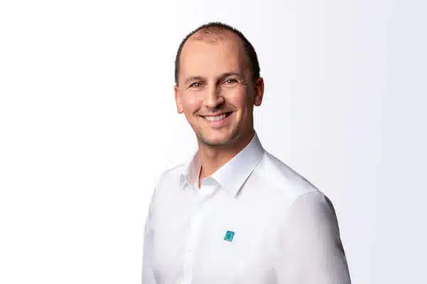 Ein lächelnder Mann mit kurzen Haaren und weißem Hemd mit Knöpfen steht vor einem schlichten, hellen Hintergrund.