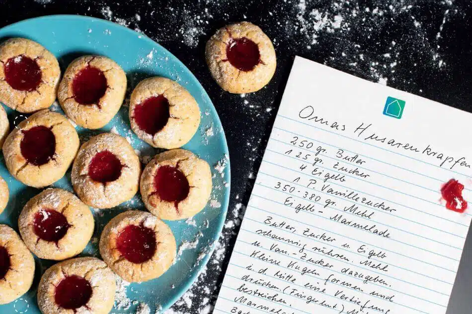 Ein blauer Teller mit Marmelade gefüllten Keksen steht auf einer gepuderten Fläche neben einem handgeschriebenen Rezept mit dem Titel Omas Husarenkrapfen auf liniertem Papier mit einem kleinen Fleck roter Marmelade.