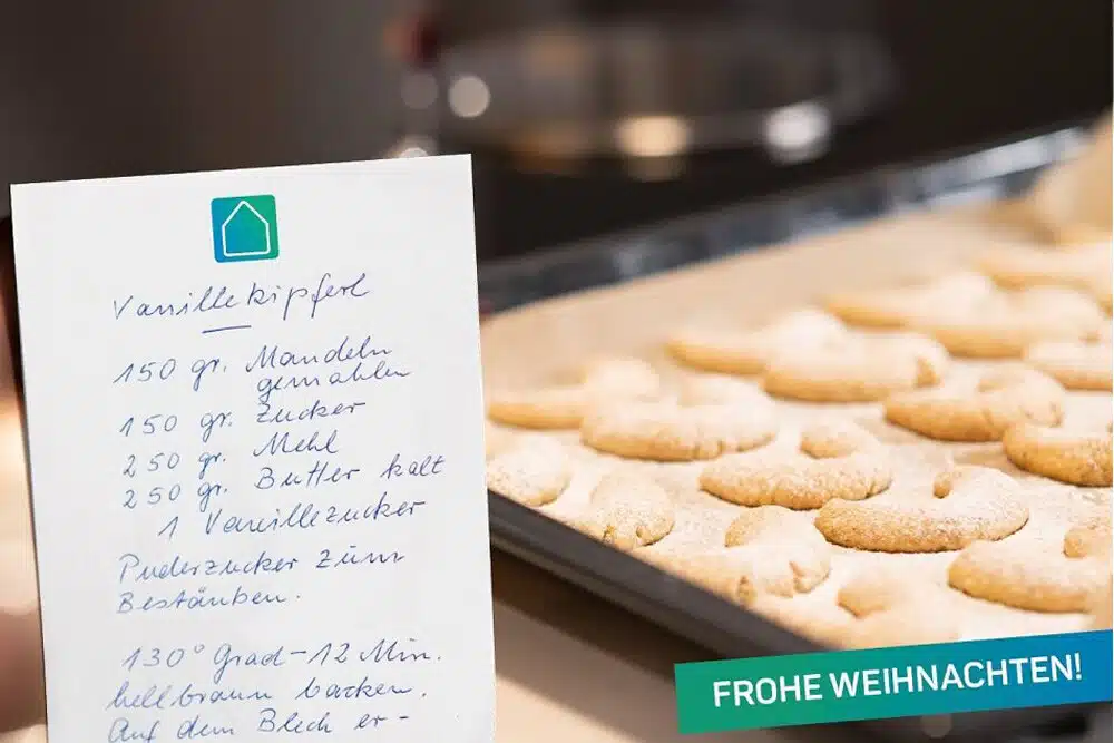 Ein handgeschriebenes Rezept für Vanillekipferl steht vor einem Backblech mit frisch gebackenen Keksen. Am unteren Rand ist ein türkisfarbenes Banner mit der Aufschrift FROHE WEIHNACHTEN! angebracht.