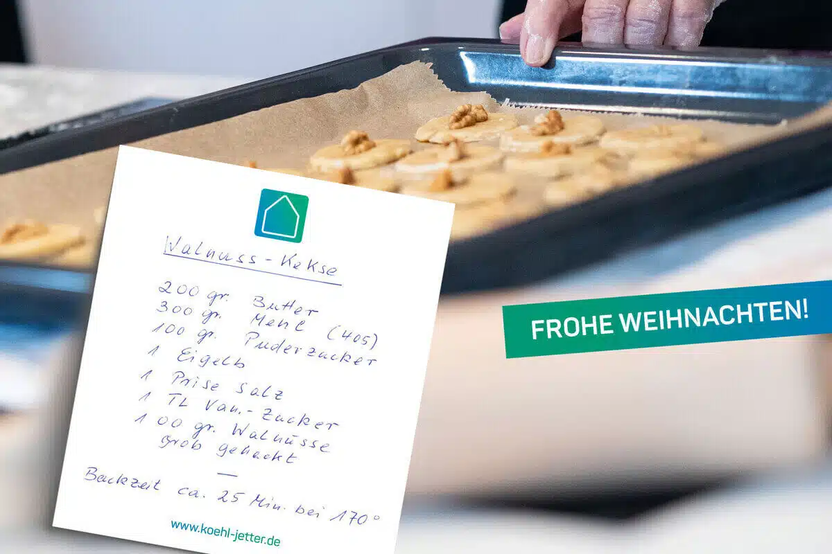 Ein handgeschriebenes Rezept für Walnusskekse in deutscher Sprache ist im Fokus, während ein Backblech mit ungebackenen Keksen und einer Hand, im Hintergrund verschwommen ist. Auf einem grünen Schild steht FROHE WEIHNACHTEN!.